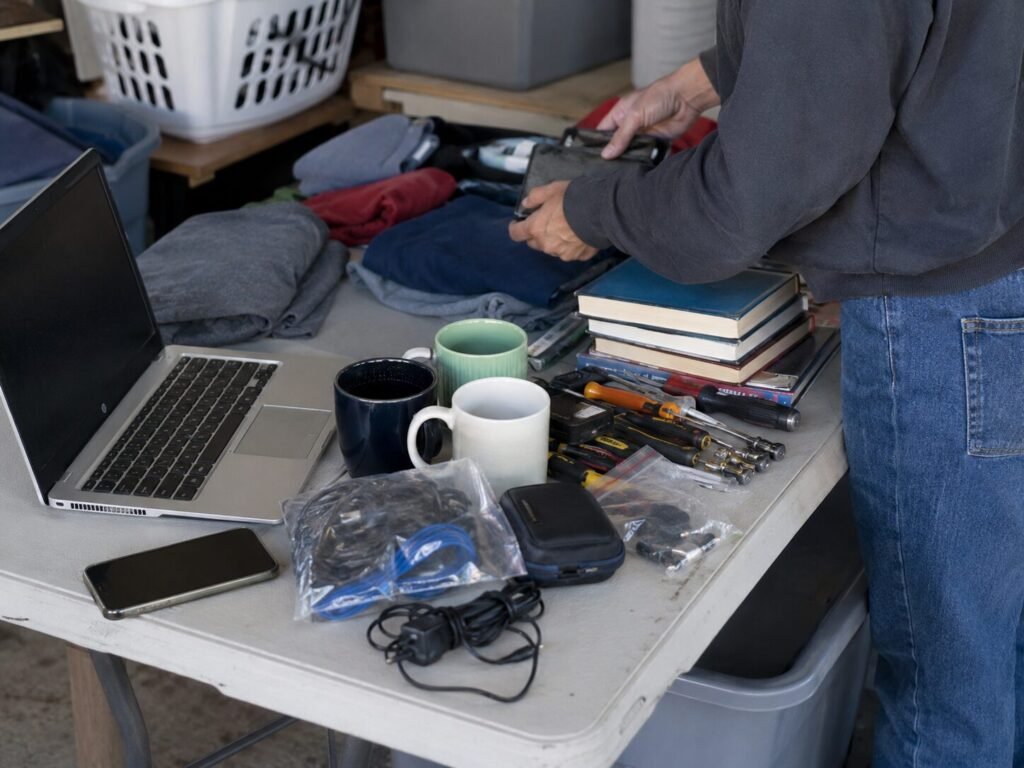 Person sorting household items, tools, books, clothing, and electronics for resale beside a laptop and phone.